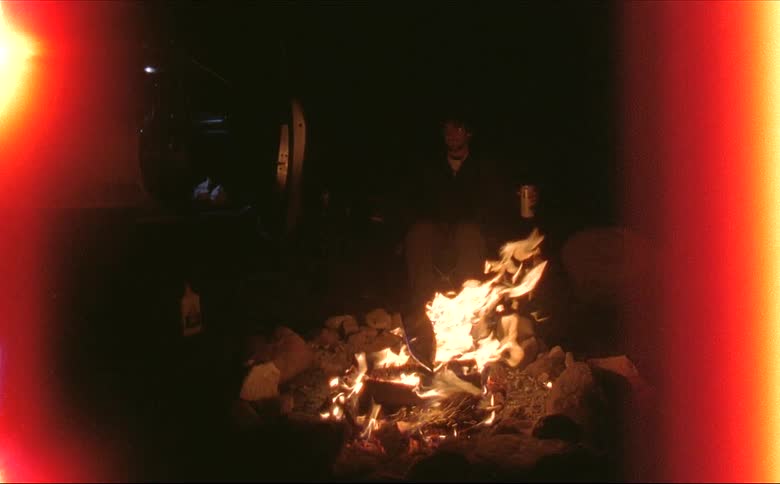A man sitting by a burning campfire at night shot on 16mm film