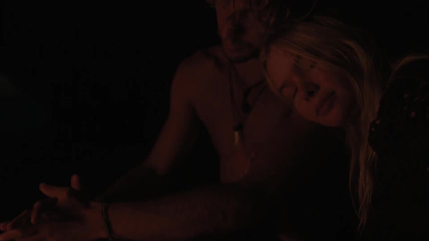 A woman rests her head on the shoulder of a man while sitting by a fire at night