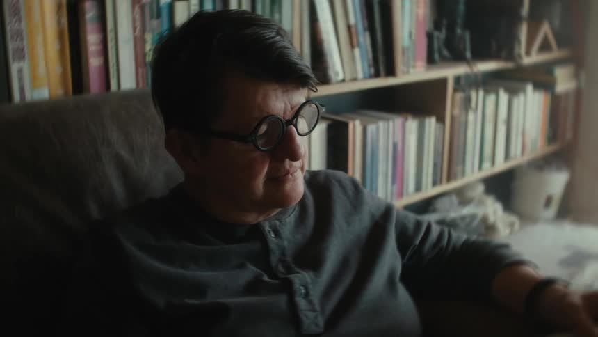 A person with short hair sitting in an armchair with books on shelves behind her