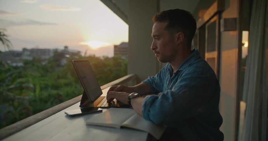 Man works on a laptop over a tall desk on the balcony of an apartment on a tropical city