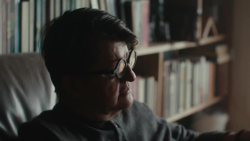 A man wearing glasses is sitting in a room with bookshelves on the wall and he is reading