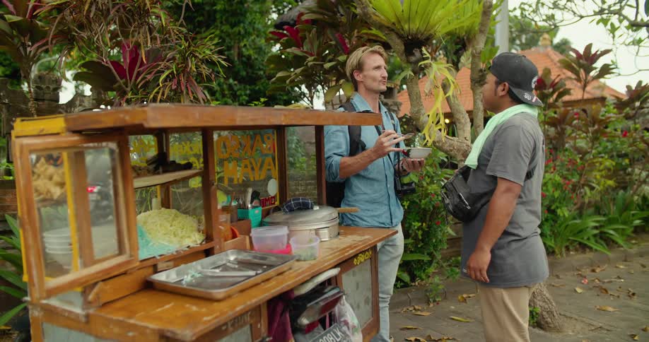 A man standing next to a food cart eating from a bowl talking to the vendor