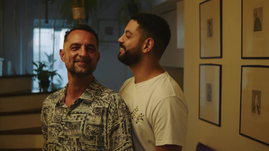 Two men stand together at home and one leans over and kisses the other