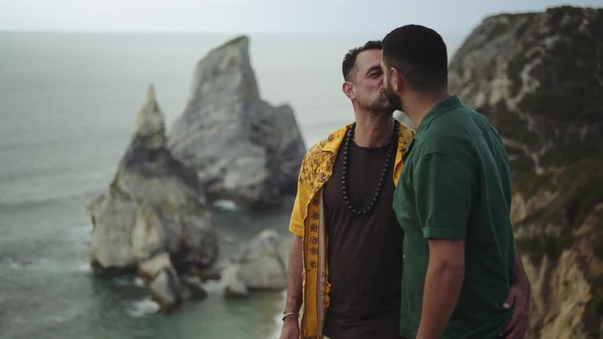 Two men affectionately share brief kisses while looking over an ocean landscape
