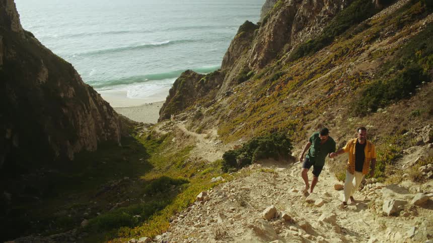 Two men holding hands while climbing up a hillside on a beach
