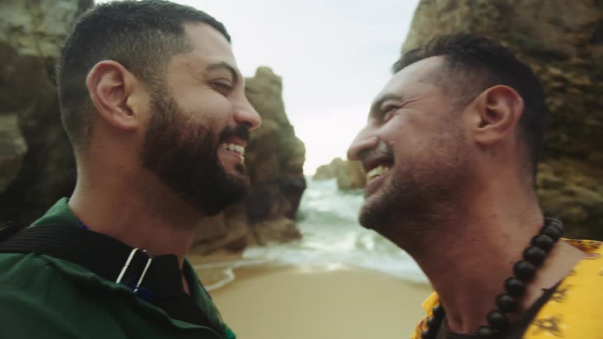 Two men laugh while kissing on the beach