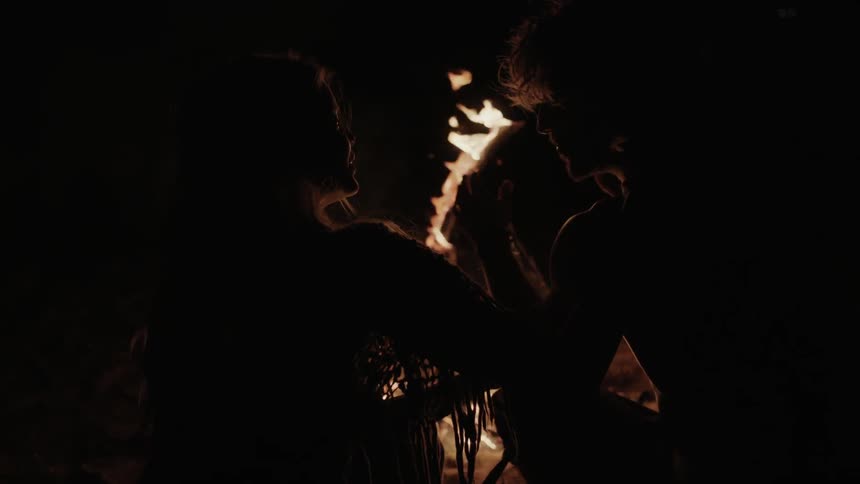 A man and woman sit laughing and warming themselves in front of a fire outdoors at night