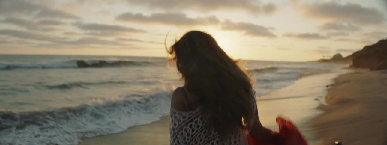 Woman walks along beach toward sun sitting on the horizon on a windy day