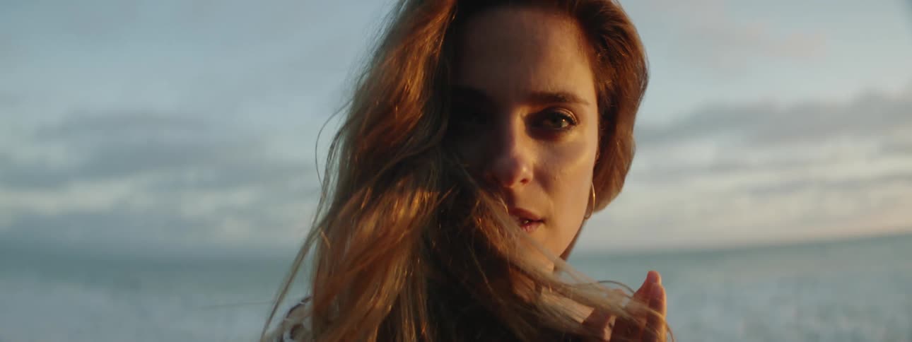 A woman is standing on a beach and putting her fingers through her hair as it moves in the breeze