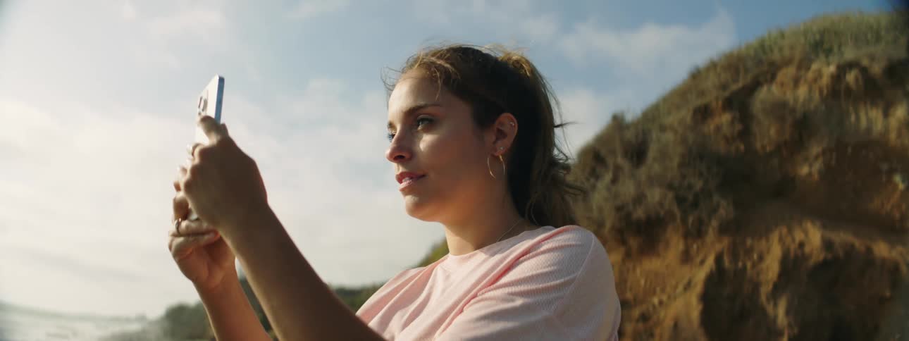 Woman on beach smiles while taking photos with her smartphone