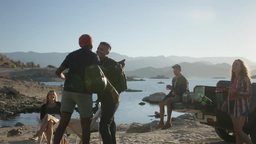 A small group of people are partying on the side of a small lake and two guys shake hands and share a friendly hug