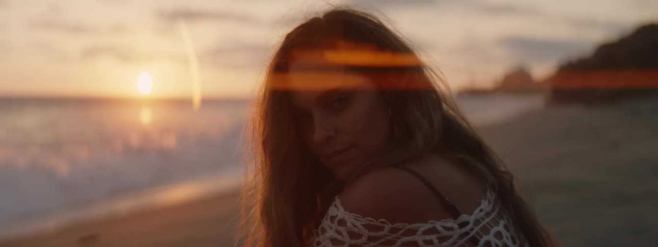 A woman is standing on a beach and she turns her head as waves roll in to shore and the sun is on the horizon