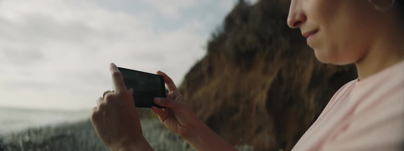A woman standing outdoors uses her smartphone to film the coastline