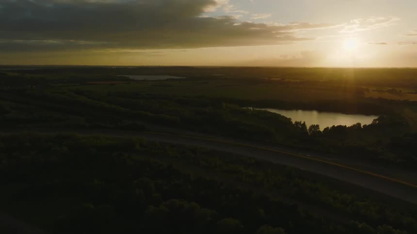 A road lined with trees bends near a lake and the sun is shining brightly on the horizon
