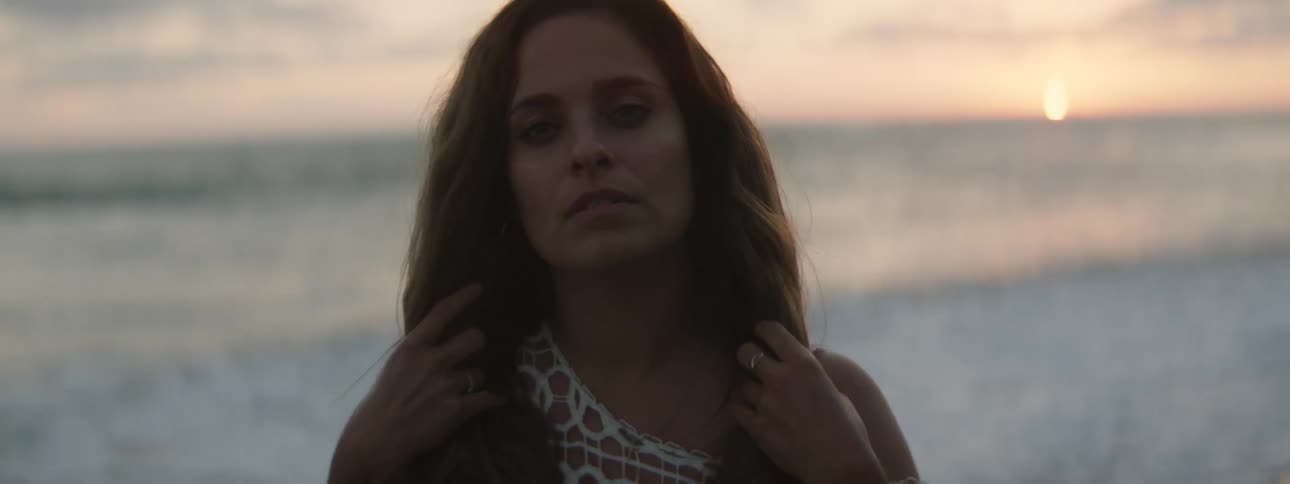 A woman is standing on a beach and moving her hands over her hair as waves roll in to shore and the sun is on the horizon