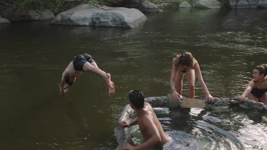 A man dives into a river as his friends sit in a hot tub and watch