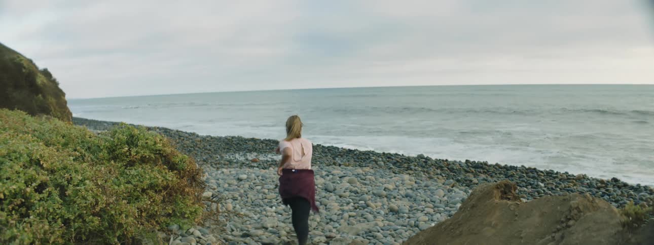 A woman walks over rocks and onto the stone covered shoreline as waves roll in under a cloudy sky