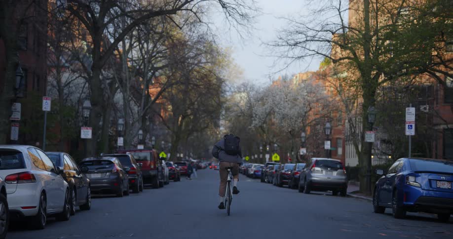 Man with dark blue jacket brown pants and black backpack rides bicycle on road next to cars parked on the street
