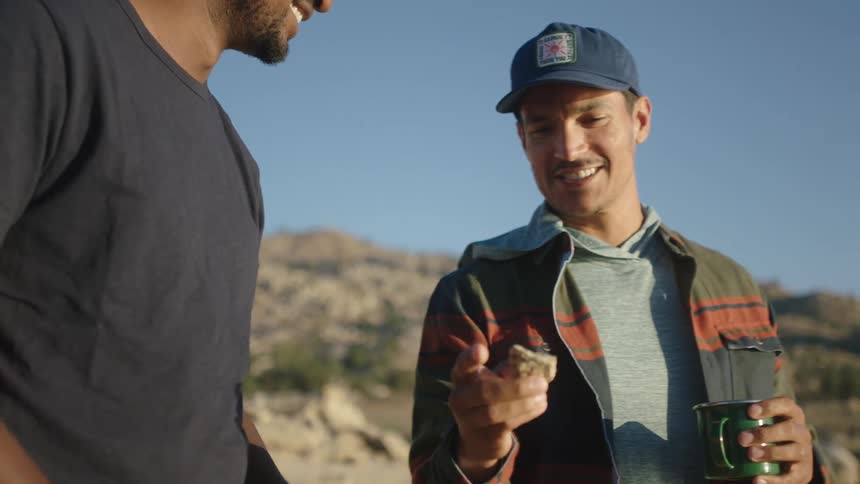 Two men are standing outside and one man is holding a rock showing it to the other man as they talk and there are mountains in the distance