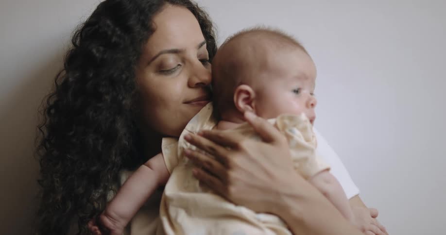 A woman is leaning against a wall holding her baby and patting their back