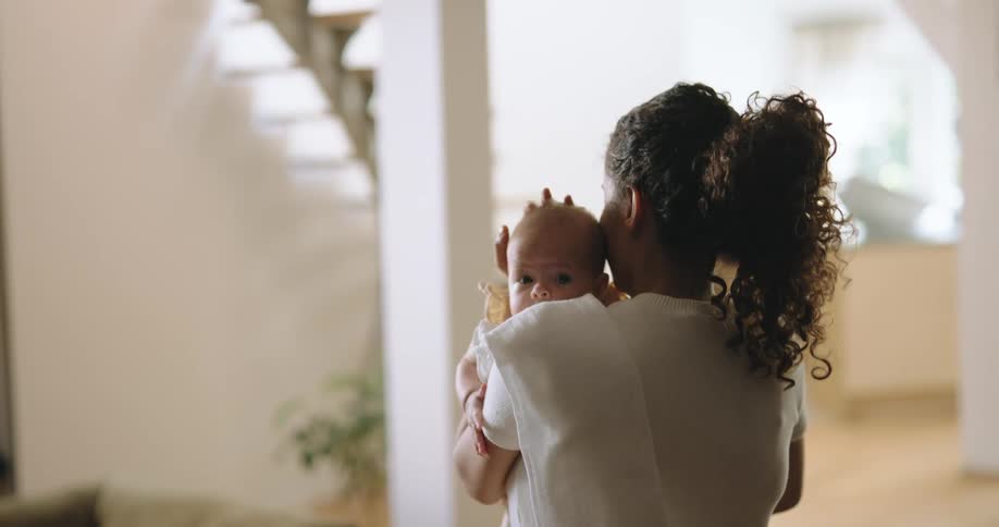 Mother with curly hair and white shirt holds newborn baby in living room at home
