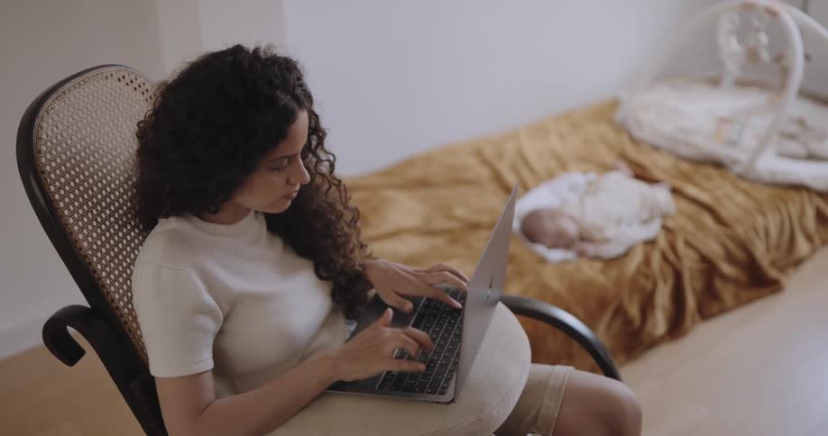 Young mother sitting on a rocking chair works on a laptop while keeping an eye on a baby