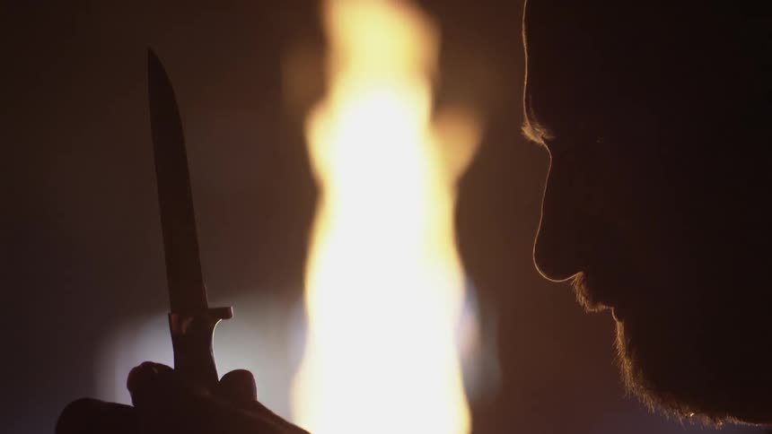 A man examines a knife blade beside a large flame