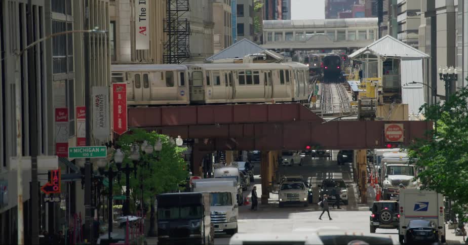 Pedestrians and vehicles move under elevated tracks as two trains move through downtown