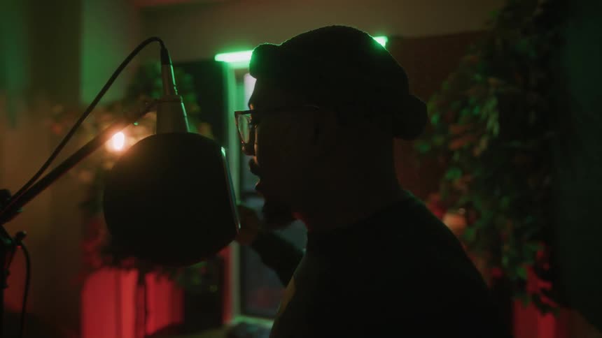 Man partially silhouetted against window in colorfully lit room records with a large microphone