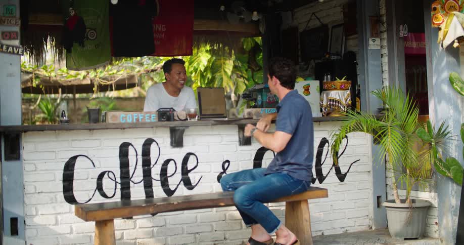 Male customer sits at outdoor bench at caf talking to the proprietor as car drives by