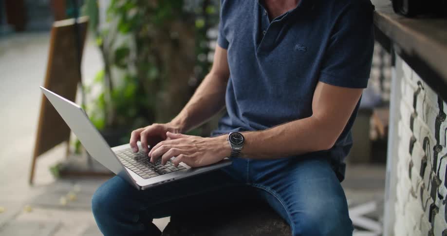 Man with blue polo shirt and jeans sits at bar and types on laptop