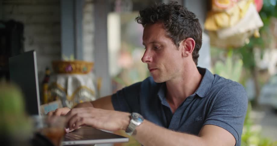A man takes a sip of his drink while thinking and working on a laptop in an open air cafe