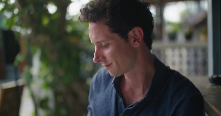 A man smiles as he sits outside on a porch typing on a laptop balanced on his leg