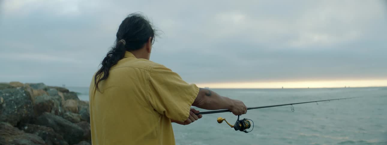 Man with ponytail and yellow shirt casts fishing pole on the coast