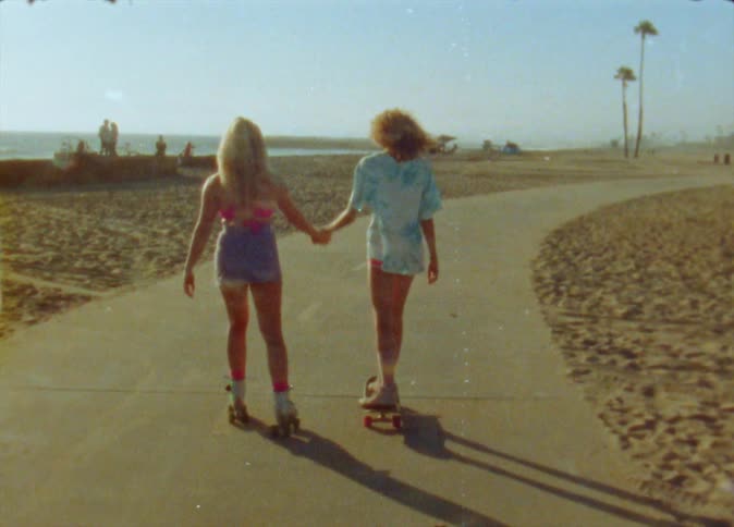 A girl on roller skates and a girl riding a skateboard are holding hands as they travel on a road near the beach as people are standing in the distance