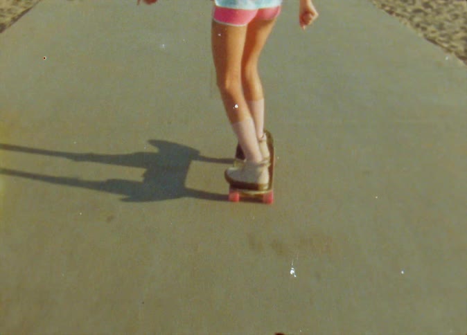 A little girl is riding a skateboard on a road to the beach and another girl is roller skating ahead of her as people walk on the road