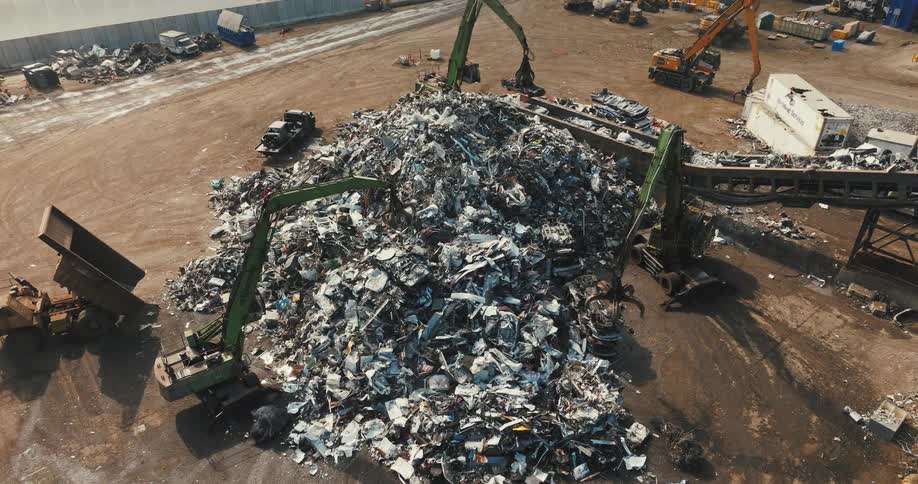 Machines with claws are moving metal scraps to a conveyor belt leading to a compressor to be compacted