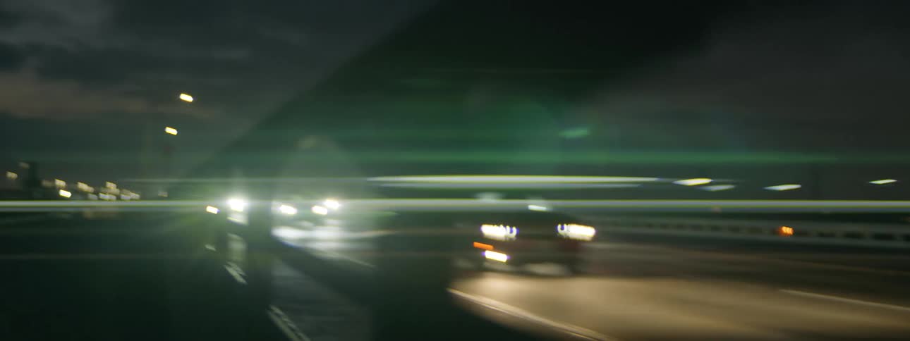 Headlights shine in the darkness as traffic moves across a highway bridge and lights from tall city buildings are in the distance