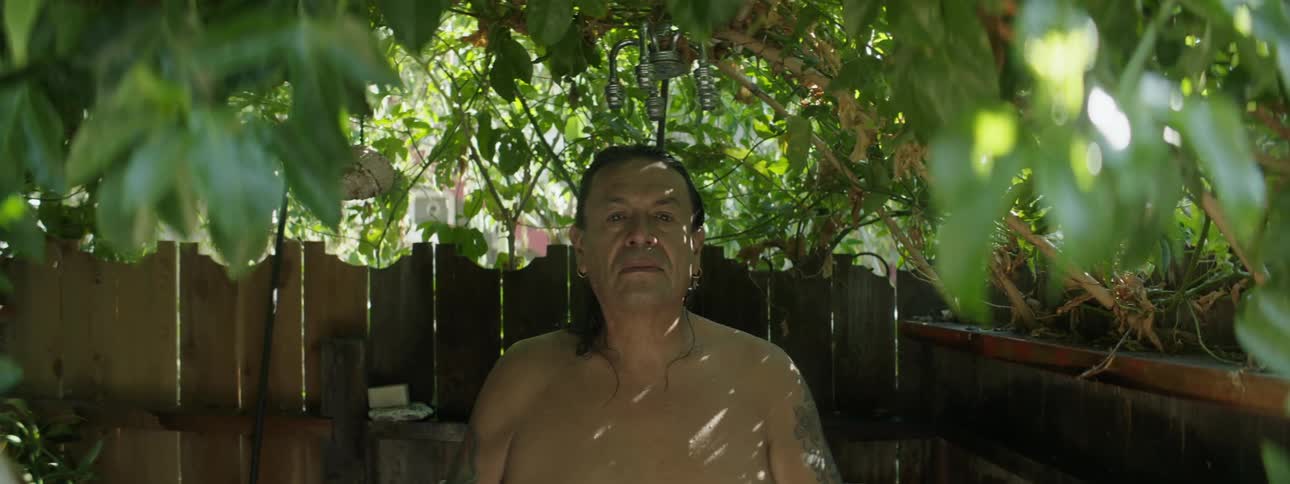 A shirtless man looks into the camera while standing beneath the showerhead of an outdoor shower