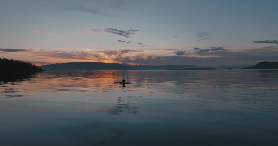 Person paddles across reflecting water as mountains on the distant horizon are silhouetted against the red sky