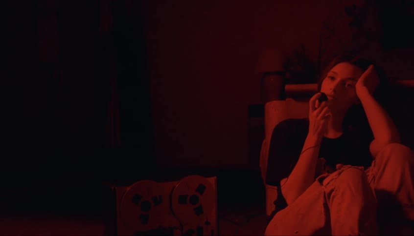 A woman is sitting in a chair in a room filled with red light and an old reel to reel video player