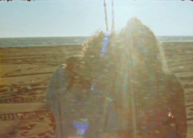 Two female friends stand in side hug and face the ocean on bright sunny beach