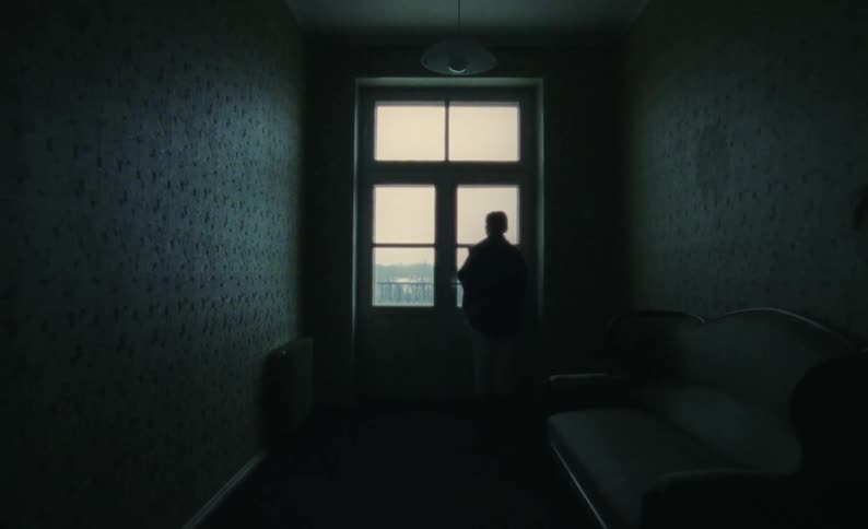 Woman silhouetted against light approaches windows and touches glass while looking out in dim room