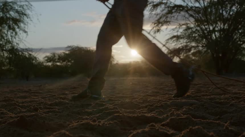 A horses legs and hooves kick up dirt as it walks followed by a cowboy in boots and spurs holding a rope