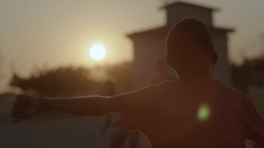 A small group of boys are playing in front of a house and a ball falls from the sky and one of the boys kicks it as the sun is shining brightly on the horizon