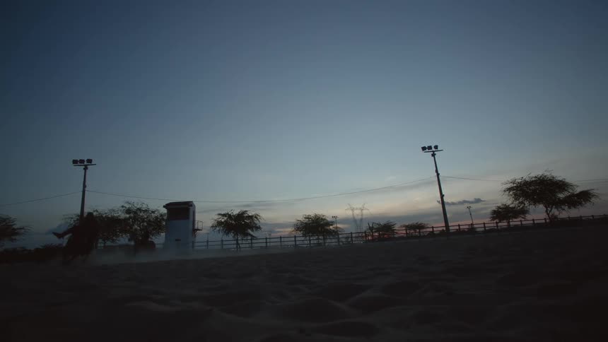 A pair of people ride horses around large sandy corral at sunset