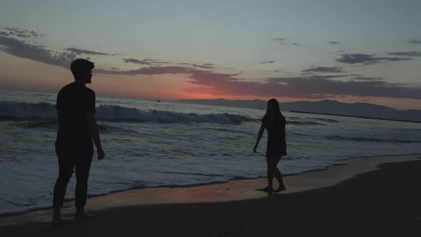 The silhouette of a man and woman standing barefoot on the wet sand at sunset