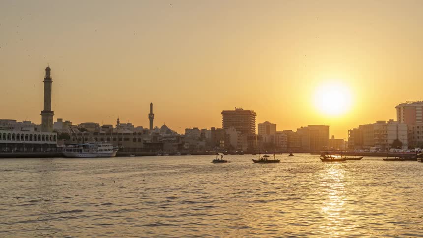 The sun shining golden over the city buildings and dubai creek river with boats traveling
