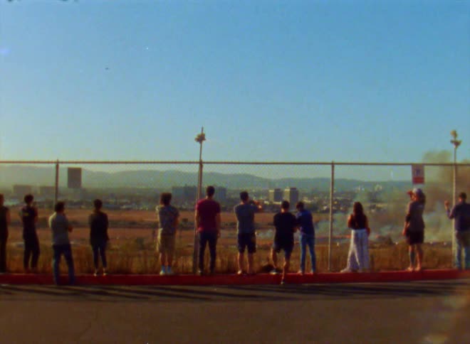 Several people are standing along a fence looking at something burning as smoke rises on the other side of the fence