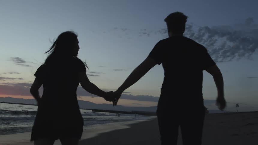 Man and woman holding hands running on the beach next to ocean are silhouetted against the sky